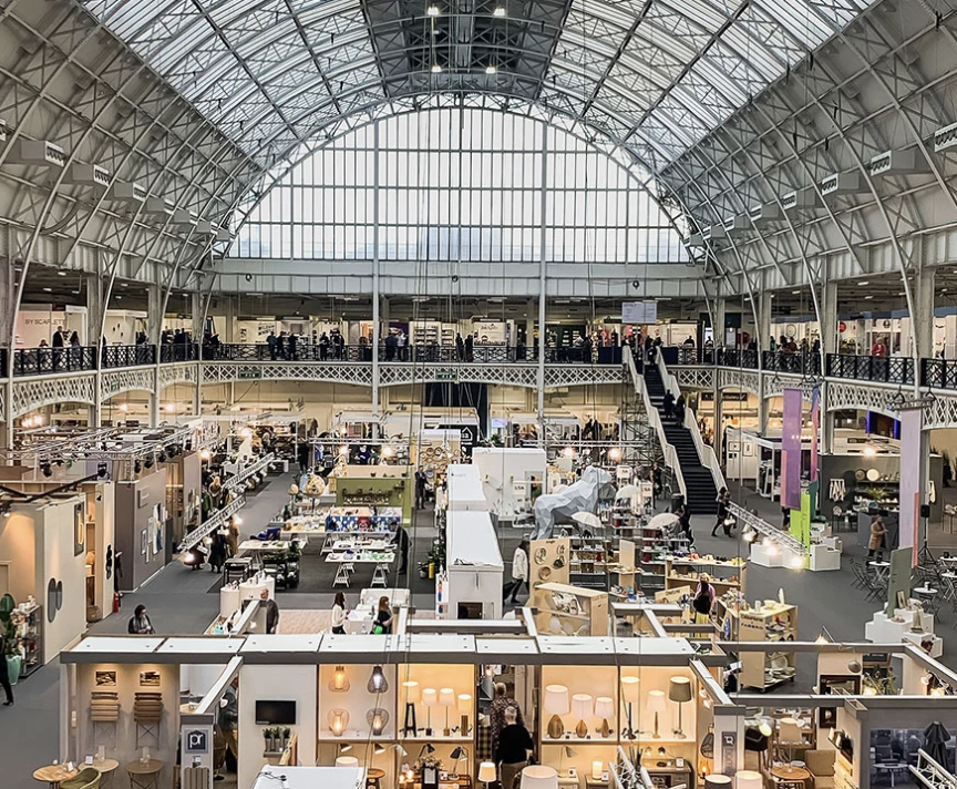 A wide view of a busy trade show hall under the vaulted glass roof at Olympia London, with rows of exhibitor stands lit up below.