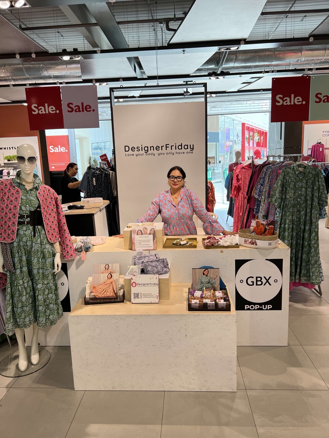The DesignerFriday popup stand set among the sale rails at a John Lewis GBX pop-up, with block-print dresses on display either side of the branded stand.