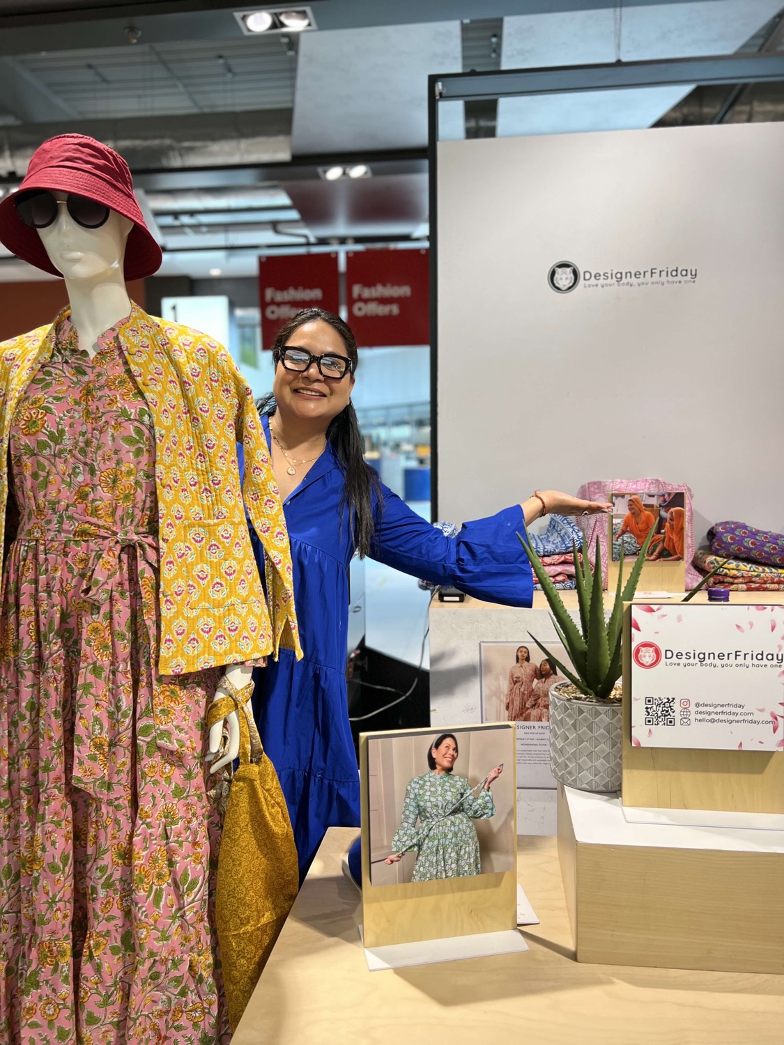 Bhavna Rishi standing beside a mannequin in a yellow and pink block-print dress, gesturing towards the DesignerFriday products as she tells the brand story.