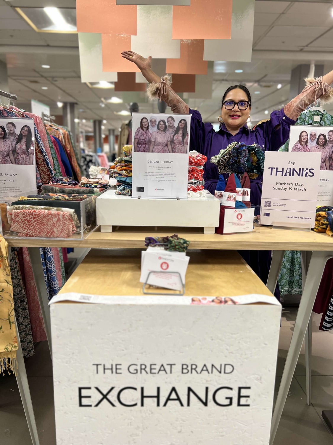 Bhavna Rishi in a purple outfit with arms raised behind the DesignerFriday stand at The Great Brand Exchange, with a Mother's Day thank you sign on the table.