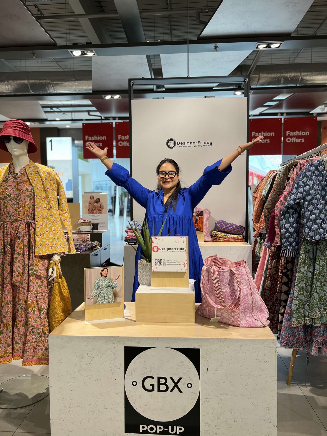Bhavna Rishi standing arms outstretched behind the DesignerFriday popup stand at a John Lewis GBX pop-up, surrounded by block-print dresses and accessories.